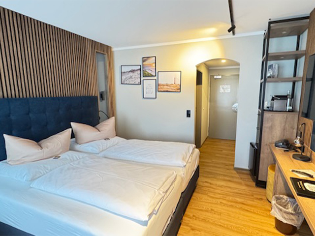 Doppelzimmer im Hotel Ennen, links befindet sich ein Boxspringbett, daneben ist der Zimmereingang und rechts ist ein Regal mit Schreibtisch erkennbar.