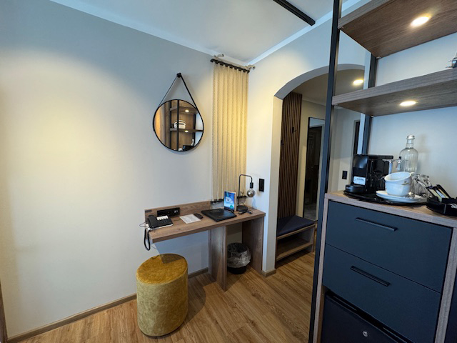 Blick in ein Doppelzimmer im Hotel Ennen, rechts befindet sich ein Regal in dem Wasser und eine Kaffeemaschine steht, dahinter ist ein Schreibtisch sowie eine Garderobe zu sehen.