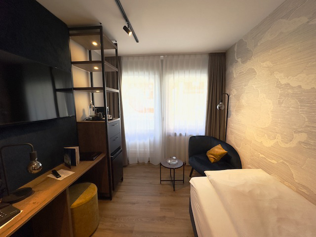 Einzelzimmer im Hotel Ennen, Blick in Richtung Fenster. Rechts im Bild befindet sich das Bett sowie ein Sessel. Auf der linken Seite ist ein Regal, ein Fernseher sowie ein Tisch zu sehen.